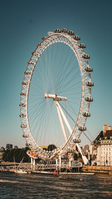 A large observation wheel, known as the London Eye, situated along the River Thames with a clear blue sky background. The wheel features multiple enclosed glass capsules evenly spaced along its rim, s