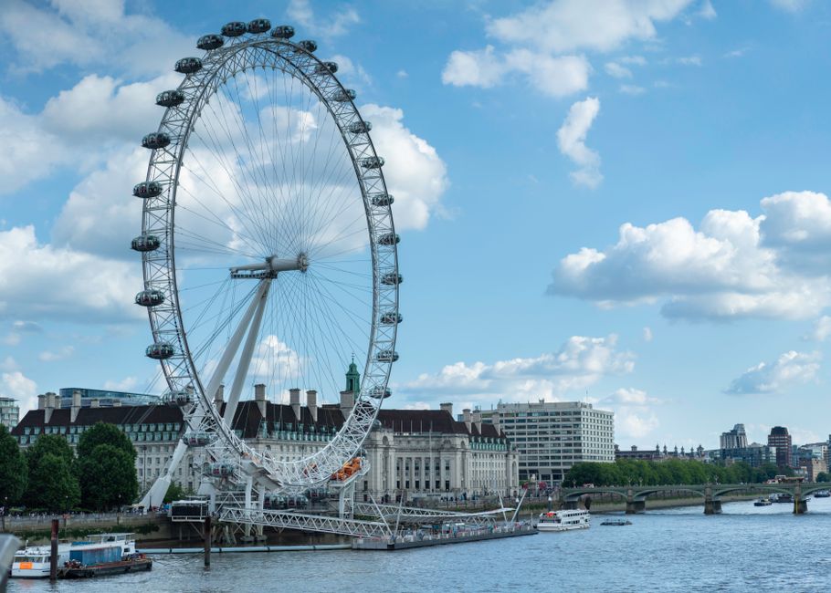 A large observation wheel, known as the London Eye, situated along the River Thames with a clear blue sky background. The wheel features multiple enclosed glass capsules evenly spaced along its rim, s